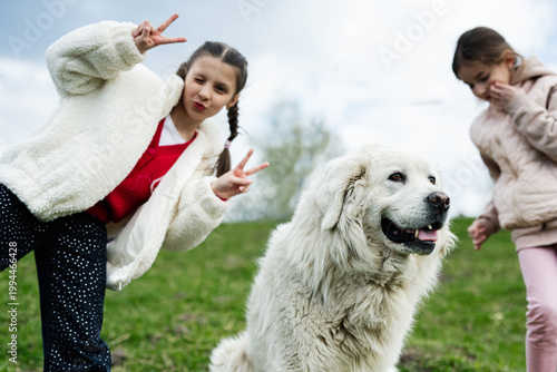 Two young girls playing outdoors with a large white dog on a grassy hill, happy pet friendship and playful spring moment