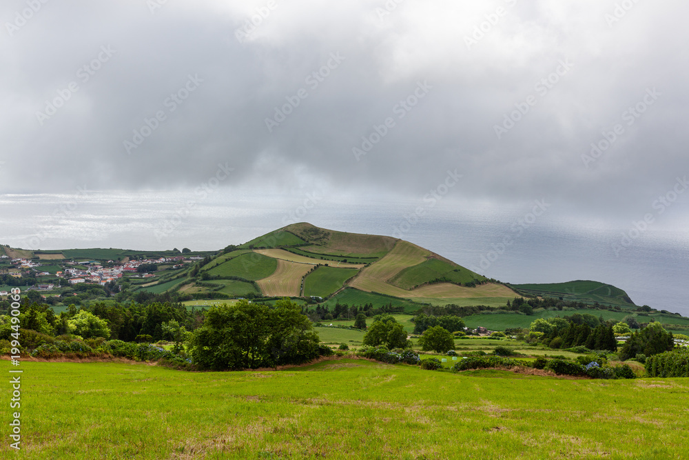 Fototapeta premium Atlantic View, San Miguel Island, Azores, Portugal. Rolling green hills with fields under cloudy sky. Distant view of coastal landscape and ocean. Small town nestled in the valley.