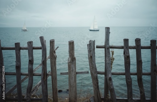 Weathered wooden fence stands by calm ocean water. Sailboats glide on the horizon under an overcast sky. Scene suggests coastal tranquility and journey.