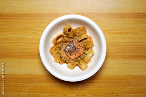 Top view of salmon pasta on white plate over wooden table, creamy sauce coating wide noodles with grilled fish. Minimal food styling, warm tones, gourmet Italian cuisine concept, restaurant menu.