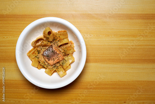Top view of salmon pasta on white plate over wooden table, creamy sauce coating wide noodles with grilled fish. Minimal food styling, warm tones, gourmet Italian cuisine concept, restaurant menu.