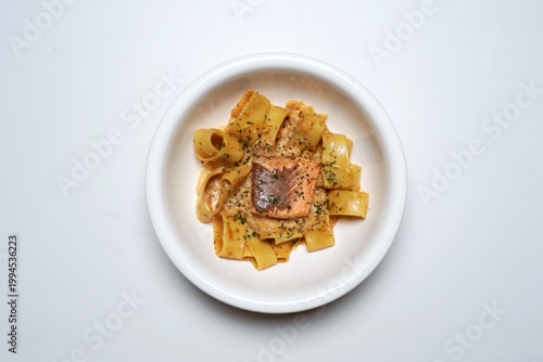 Overhead shot of creamy salmon pasta served on white dish with clean white background, wide noodles and seared fish detail. Modern minimal food photography, fine dining concept, appetizing meal.