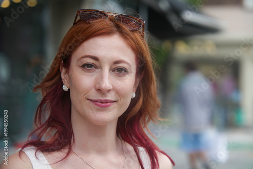 Natural portrait of a confident smiling redhead woman looking directly at camera in an outdoor urban setting. Soft expression, real lifestyle moment with blurred city background and copy space.