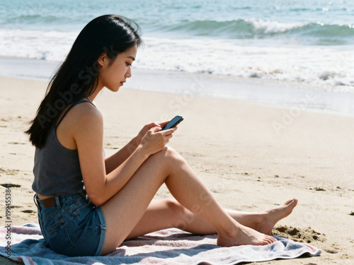 Young Asian woman sitting on beach towel by shoreline while checking smartphone