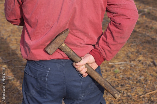 a one man criminal in red blue clothes holds a large brown hammer in his hand behind his back on the street