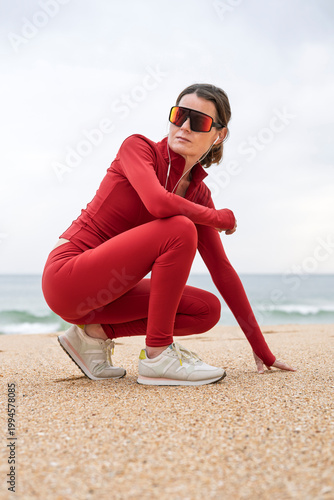Fit Woman in Red Sportswear Squatting on Beach With Sunglasses and Earbuds