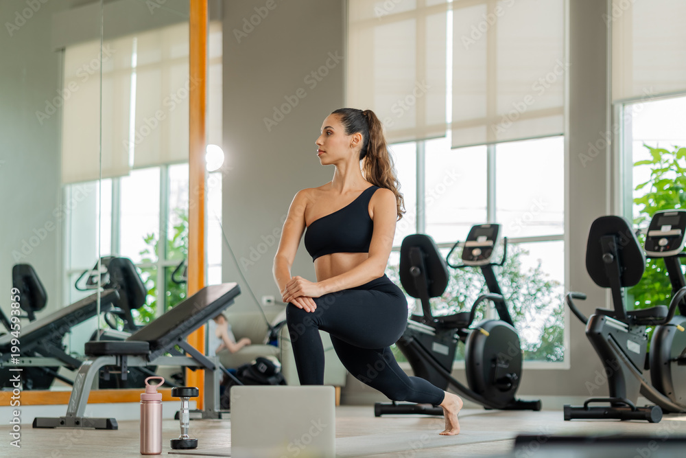 Fototapeta premium Athletic woman performs lunge exercise near mirror in well-lit gym. Female athlete doing yoga and fitness exercises with virtual class on computer, digital health and active lifestyle concept.