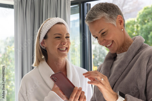 Female mother and daughter smiling, wearing bathrobes, holding smartphone near glass doors at home