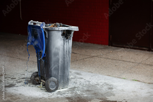 Burnt and melted plastic trash can on a city street after a fire, showing severe heat damage, soot stains, and urban decay
