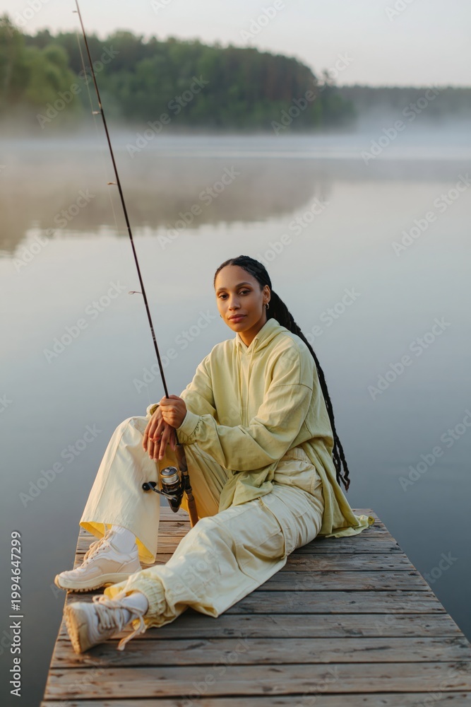 Fototapeta premium Woman sitting with fishing rod on misty lake dock at dawn