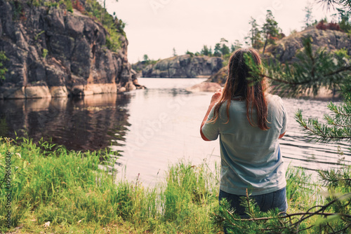 A young woman stands amidst green grass and pine branches, looking out over a tranquil bay framed by steep granite cliffs and a forested northern shoreline.