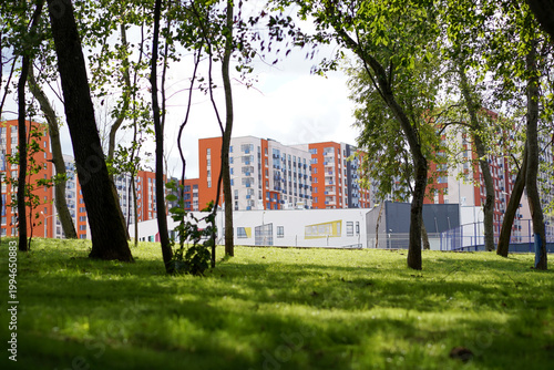 A view of vibrant, modern orange and white apartment buildings and a contemporary school facility as seen through the lush green trees of a nearby park.