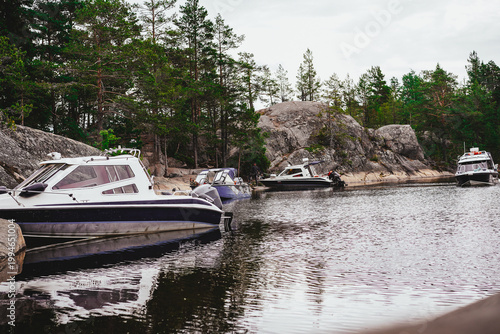 Several modern motorboats are moored along a rugged, rocky shoreline nestled within a dense pine forest in a northern wilderness setting.