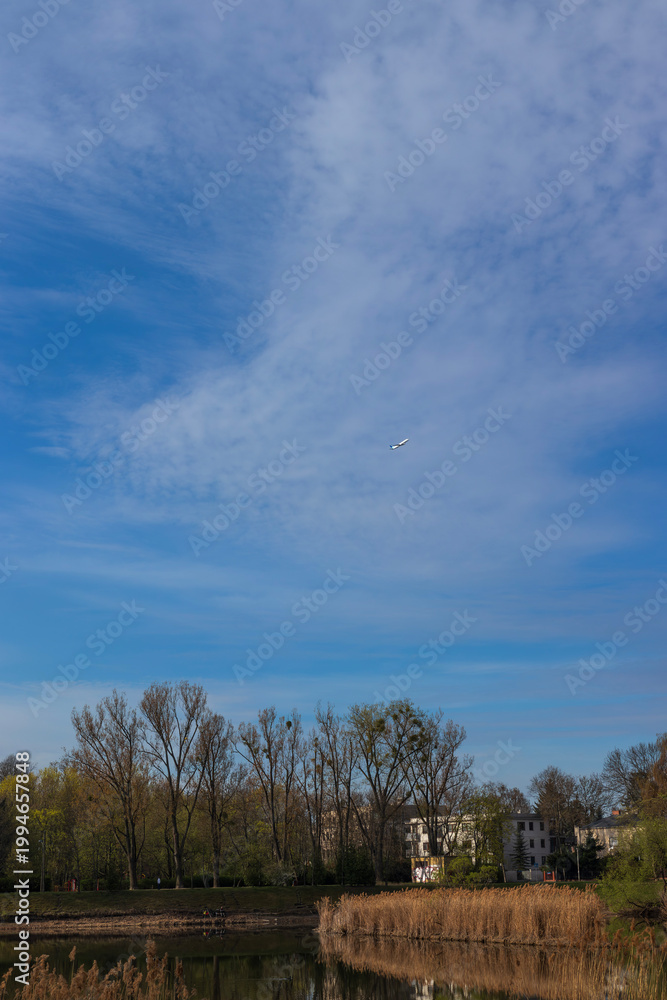 Obraz premium Airplane high in the sky above a calm lake, reeds and spring trees with dramatic clouds.