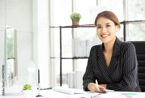 Portrait of executive financial businesswoman sitting in front of digital computer at her desk in modern office while looking at camera and smiling.