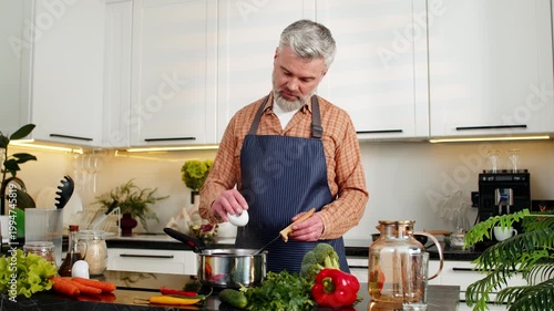 Middle-aged man cooks soup on electric stove seasoning and stirring for healthy diet in home kitchen. Mature guy feels happy while preparing homemade dinner for weight loss goals and daily wellness.
