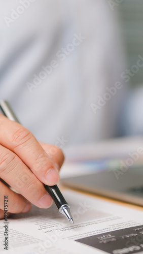 Detailed view showing male hand holding black pen writing signature paper document near credit card laptop computer inside bright office workspace professional business executive reviewing financial.