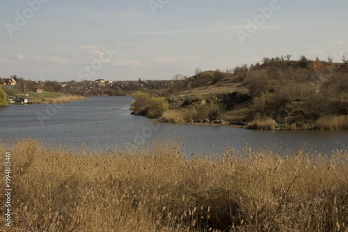 Calm river flowing through reeds and hills with rural houses in the background under a clear sky.