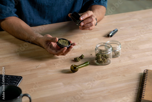 Man using cannabis grinder, with cannabis equipment and accessories on the table.
