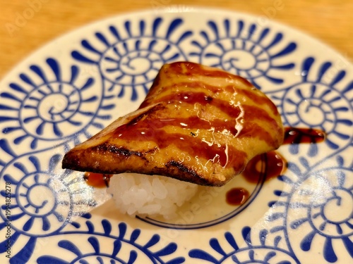 High angle close-up of a foie gras nigiri sushi with rich glaze, served on patterned ceramic plate