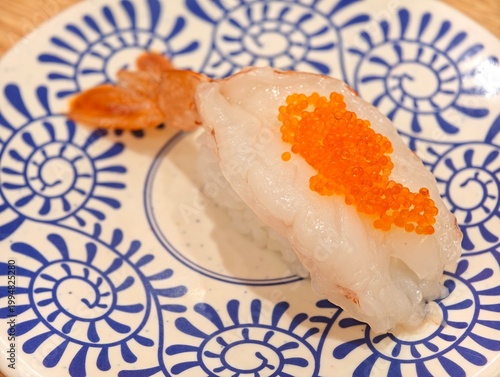 High Angle of a raw shrimp nigiri sushi topped with bright orange tobiko, served on a patterned ceramic plate