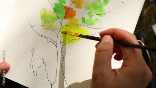A hand paints a vibrant watercolor tree with green, yellow, and orange hues on white paper using a fine brush.