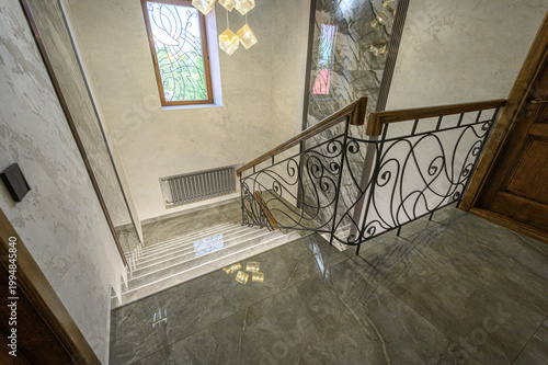 Elegant marble staircase with ornate wrought iron railing, textured walls, stained glass window, modern chandelier, polished floor, and wooden door. Luxurious, refined interior