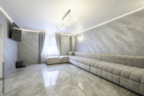 Modern minimalist living room with marble-finish floor and walls, oversized beige sofa, LED ceiling lighting, wall-mounted TV, sheer curtains, sleek design, airy ambiance