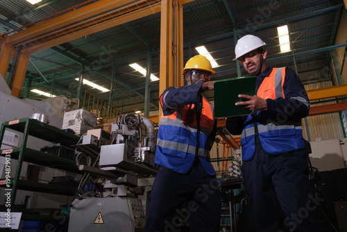 Billede på lærred Two workers review information on a tablet while standing in a large factory dur