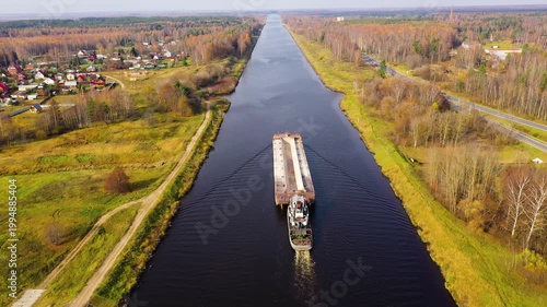 River tug with a barge. River channel for freight transportation, top view. Beautiful landscape with a wide river and autumn forest.