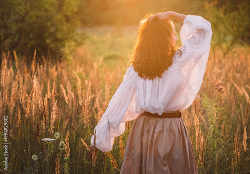 Obraz na płótnie free-spirited woman in boho outfit walking away through golden meadow at sunset