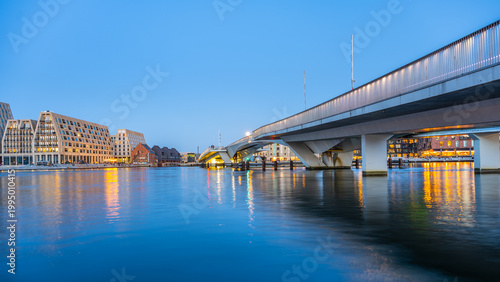 Copenhagen's Inner Harbour Bridge stands over the water as buildings reflect in the calm surface. The sky is darkening, creating a scene of city life near the waterfront.