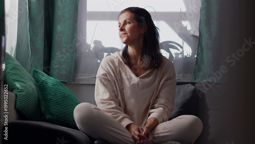 Fotografi Middle aged woman sitting cross legged on sofa by window in soft daylight appear
