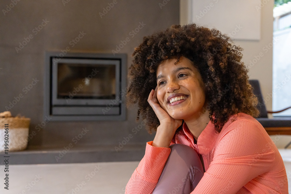 Naklejka premium African American woman sitting on floor by fireplace wearing coral top and mauve leggings, smiling
