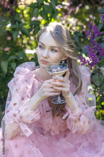A portrait of a young woman with long hair in a pink dress among blooming lilacs. She sits with a glass of wine, relaxing on a picnic. Spring blossoms.