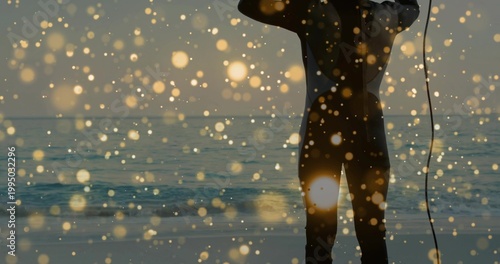 Standing silhouetted surfer wearing dark wetsuit facing ocean at shoreline, holding surfboard leash