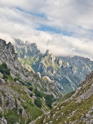 Rugged Limestone Peaks in Picos de Europa - Majestic and dramatic mountain landscape with jagged limestone peaks, green slopes, and clouds hanging low in Picos de Europa, Asturias, Spain.