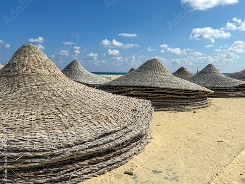 Several round woven straw beach umbrellas sit on golden sand against a clear blue sky, peaceful tropical resort, empty summer beach, holiday relaxation.