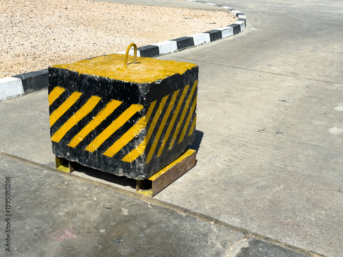 A square concrete barrier with black and yellow warning stripes sits on an asphalt road near a curb. Industrial safety, road construction, urban infrastructure.