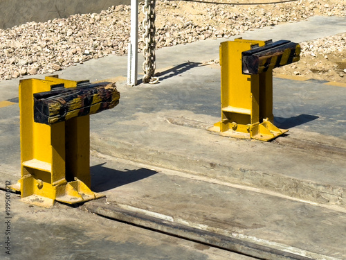 Two bright yellow metal safety bollards with black and yellow rubber bumpers are bolted to an outdoor concrete surface. Industrial safety, facility protection, loading zone.
