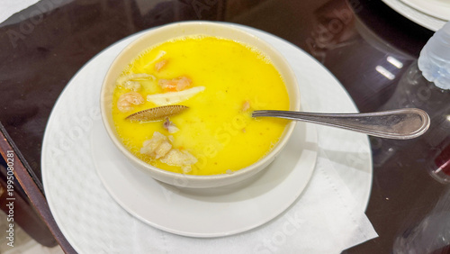 A warm bowl of yellow savory soup rests on a white plate with a spoon, cozy homemade cooking, healthy hot meal.