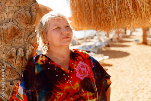 Relaxed senior woman leaning on palm tree at tropical beach resort. Peaceful retirement, mental health and body positivity concept.