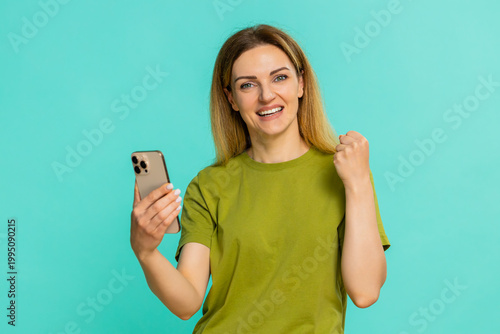Young woman smiling while holding smartphone, proudly showing achievement or celebrating victory in game, receiving good happy news. Caucasian girl on blue background with joyful positive reaction.
