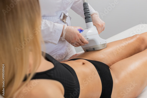 Young adult woman gets cavitation vacuum therapy on thighs by cosmetologist using LPG machine. Procedure helps reduce girl cellulite, tighten skin, and improve circulation in modern beauty clinic.