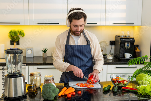 Middle-aged man chops tomatoes for fresh salad while wearing headphones singing in home kitchen. Young guy feels happy follows healthy diet for weight loss meal prep plus simple cooking for fitness.