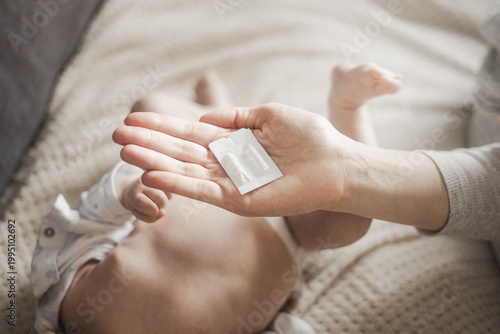 A woman holds two medical suppositories in white packaging on her open hand over a lying baby at home