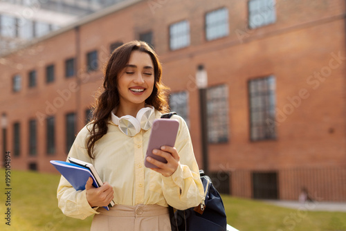 Photography Smiling lady student with headphones around her neck, looking at smartphone whil