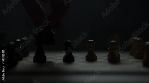 Cinematic close-up of a hand moving a chess piece in a dark atmospheric room. Low-key lighting highlights strategy, planning, and intellectual competition. Dramatic silhouette of a chess game.