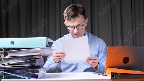 Obraz na płótnie Man in glasses intently reviewing a document at a cluttered office desk stacked