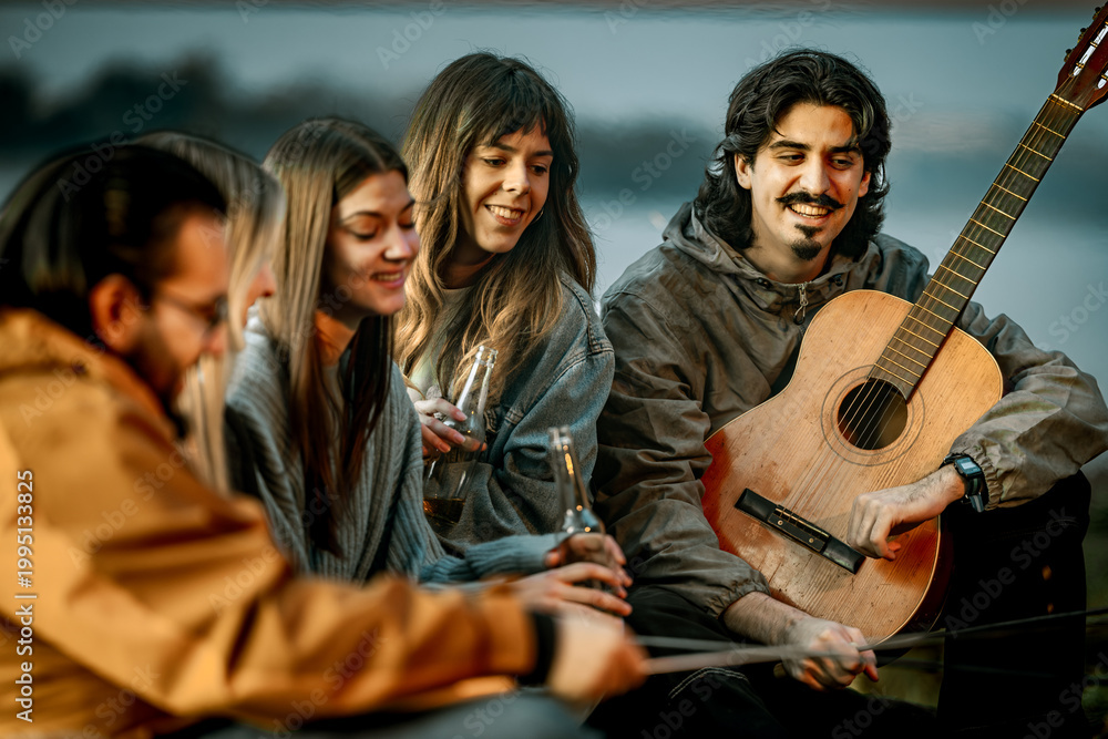 Fototapeta premium Group of friends enjoying time together near water while playing music and having drinks during sunset